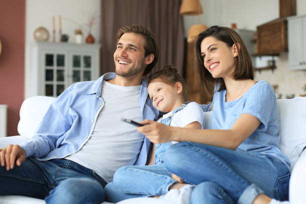 happy family with child sitting on sofa watching tv, young parents embracing daughter relaxing on couch together. - apartment television family couple imagens e fotografias de stock
