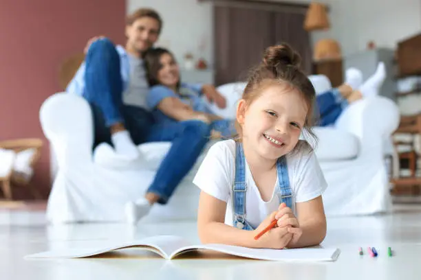 Photo of Smiling happy family sit relax on couch in living room watch little daughter drawing in album with colorful pencils. Happy weekends at home.