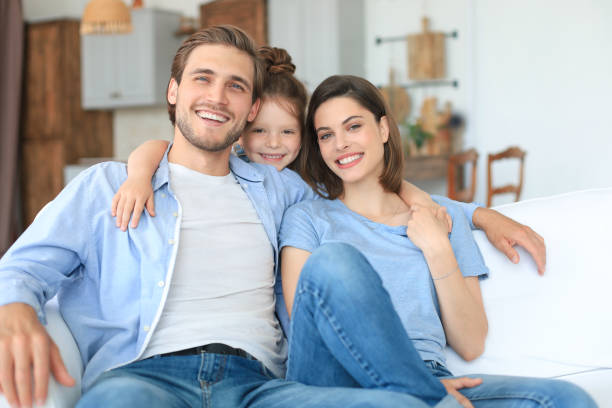 istock Positive friendly young parents with smiling little daughter sitting on sofa together while relaxing at home on weekend. 1267503380