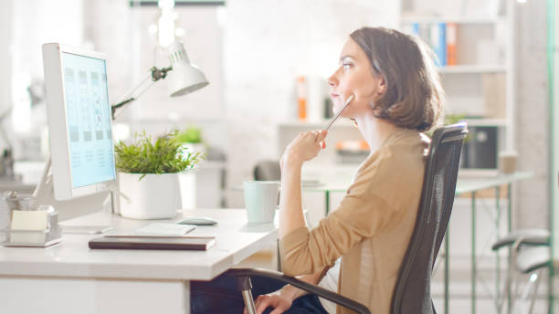 istock Focused Creative Woman Sitting at Her Desktop Computer in Her Modern Bright Office. She is Thinking about Problem Solving and Holding Her Pen. 1267506928