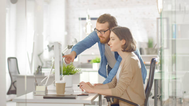 istock Young Male Manager and Beautiful Woman Discuss Working Issues at Workplace. She is Using Computer and He Stands Beside Her. Bright Modern Loft Office with Smart People Working. 1267507462