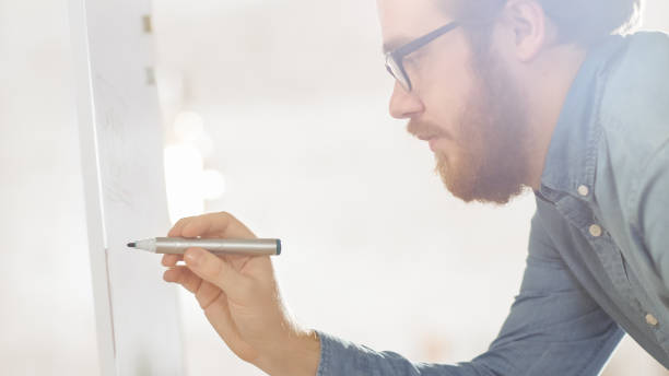 istock Creative and Successful Young Male Developer wearing Glasses and Draws Charts on a Whiteboard. Bright Modern High Tech Office with Beautiful People. 1267507486