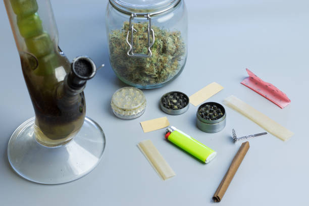 Smoking cannabis accessories and stuff on gray background. Bong, blunt and joint paper, grinder and marijuana buds in jar Smoking cannabis accessories and stuff on gray background. Bong, blunt and joint paper, grinder and marijuana buds in jar. bong photos stock pictures, royalty-free photos & images