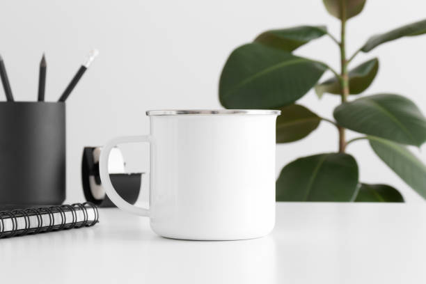 istock Enamel mug mockup with workspace accessories on a white table and a ficus plant. 1267508999