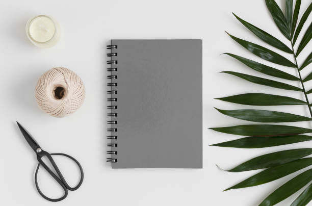 Top view of a grey notebook mockup with  workspace accessories and a palm leaf on a white table.
