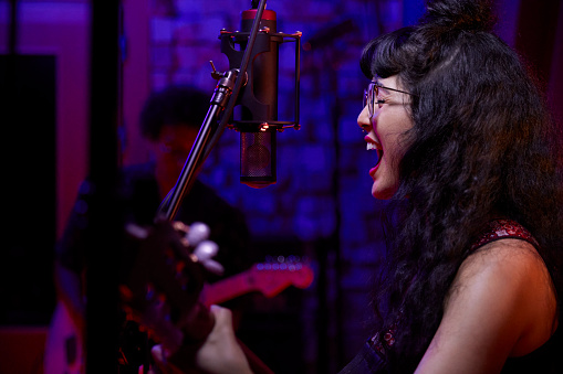 Close-up of mixed-race female singer recording vocals and playing guitar with band in professional music studio