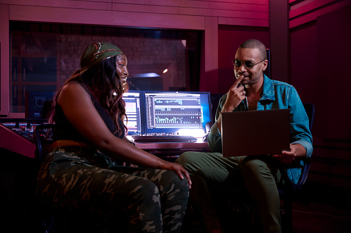 African-american female artist working on song track with male music producer on laptop in professional music studio