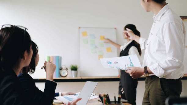 istock Businesswoman sticking adhesive notes on whiteboard while colleagues standing with arms crossed at office 1267520417