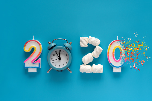 2020 year made of candles, marshmallow and alarm clock with dispersion effect on blue background. New Year 2021 celebration or end of year 2020 concept. Top view. Flat lay