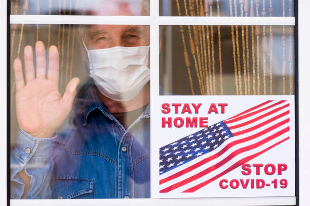 un uomo anziano dietro la finestra di casa resta a casa indossando una maschera chirurgica per evitare il contagio del coronavirus covid-19 - concetto di speranza per il futuro - isolated despair hope assistance foto e immagini stock