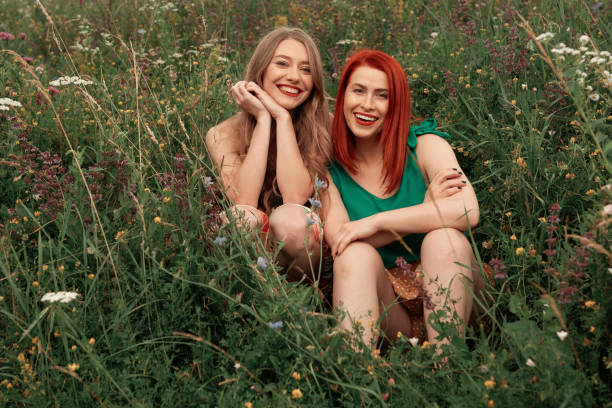 Blond best friend and redhead sitting in the meadow looking at camera with toothy smile