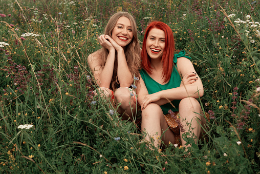 Blond best friend and redhead sitting in the meadow looking at camera with toothy smile