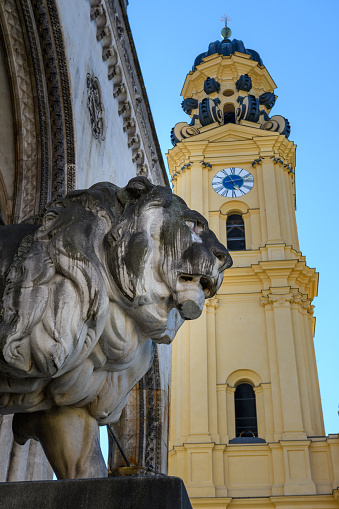 Munich, Germany: A stone lion sculpture at the Feldherrenhalle, in the background the clock tower of Theatine church at Odeonsplatz