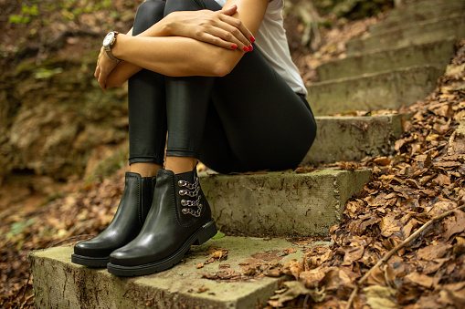 Photo of women who is sitting on the stairs outdoor surrounded by fallen autumn leaves. She is wearing modern leather black boots and casual clothes