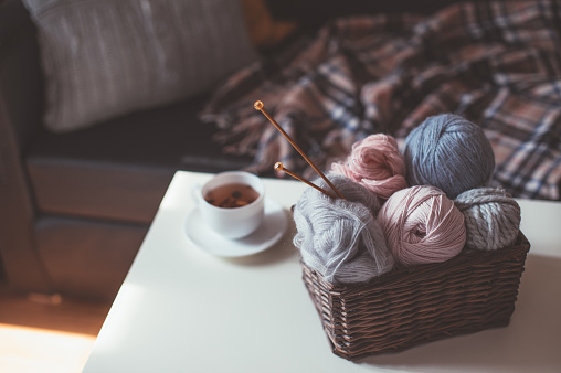 Balls of woolen yarn and knitting needles in basket and hot herbal tea in cozy interior.