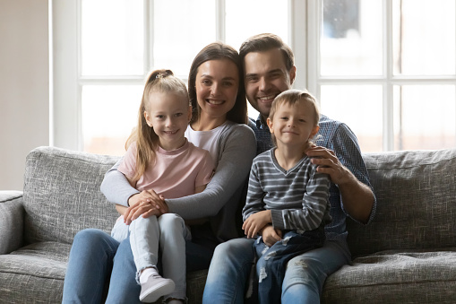 Portrait of happy young parents holding on laps little funny children, relaxing together on cozy sofa in living room. Smiling married caring couple enjoying tender loving moment with small kids.