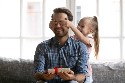 Happy little baby girl covering eyes of handsome father holding wrapped box in hands. Smiling young daddy feeling curious about surprise present, Father's Day birthday or special occasion celebration.
