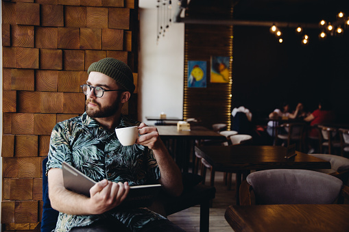 Modern fashionable male student with beard, hat and eyeglasses reading book drinking coffee and looking to the side in cafe.