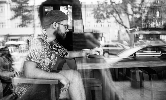 Black and white shot of modern hipster male working on laptop in cafe. Reflections.