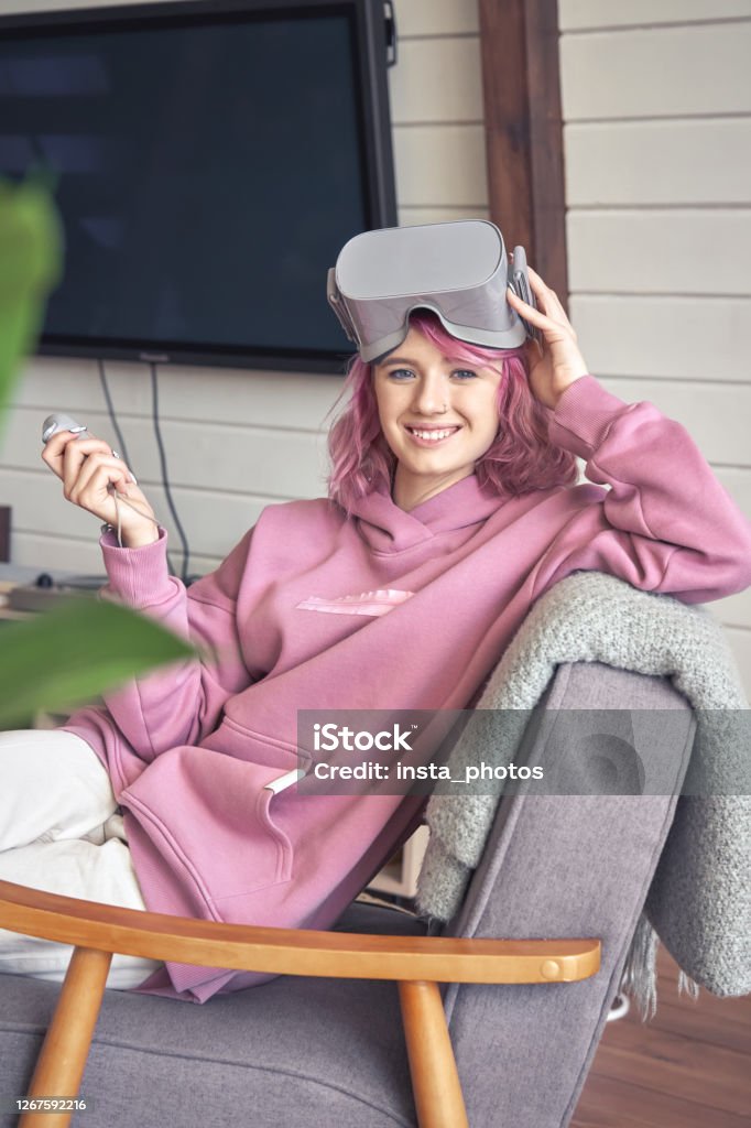 Happy hipster teen girl wear vr headset hold controller look at camera in chair. Happy hipster teen girl pink hair wear vr glasses headset hold controller sit in chair look at camera. Digital innovation video game, virtual reality 3D 360 video app entertainment. Vertical portrait Extinction Rebellion Stock Photo