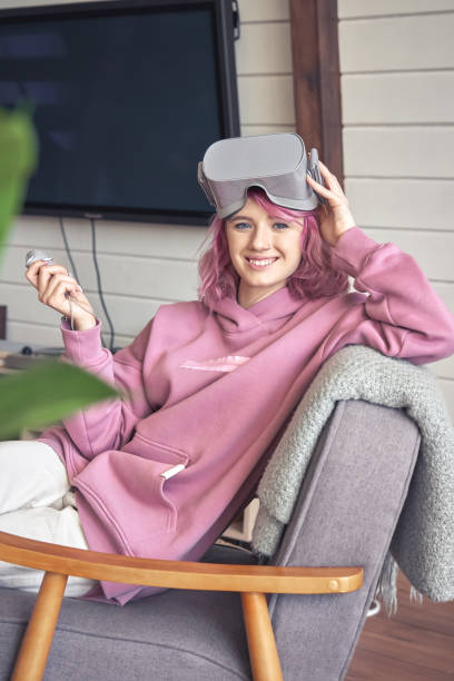 Happy hipster teen girl wear vr headset hold controller look at camera in chair. Happy hipster teen girl pink hair wear vr glasses headset hold controller sit in chair look at camera. Digital innovation video game, virtual reality 3D 360 video app entertainment. Vertical portrait extinction rebellion photos stock pictures, royalty-free photos & images