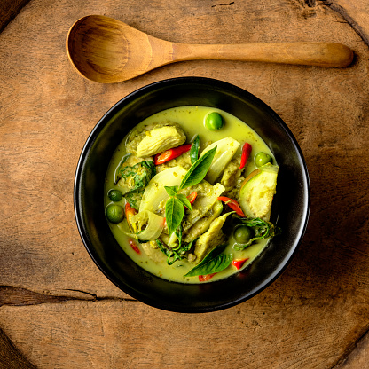 This Thai known in Thai as 'Gaeng Keow Wan Gai' dish is very famous and known all over the world and one of Thailand's popular signature dishes when it comes to Thai food. This dish consists of ingredients including a green herb-based base with fresh coconut milk, sweet basil, Thai aubergines, pea aubergines, chicken, and spicy fresh red chili. The combination of ingredients results in a sweet and spicy taste with a variety of complementary textures and is normally eaten together with fresh steamed Jasmine Thai rice or fresh rice noodles. The image was taken from directly above with the bowl and spoon set on an old worn textured wooden tray with lots of grain and character.