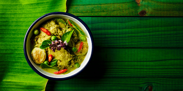 This Thai known in Thai as 'Gaeng Keow Wan Gai' dish is very famous and known all over the world and one of Thailand's popular signature dishes when it comes to Thai food. This dish consists of ingredients including a green herb-based base with fresh coconut milk, sweet basil, Thai aubergines, pea aubergines, chicken, and spicy fresh red chili. The combination of ingredients results in a sweet and spicy taste with a variety of complementary textures and is normally eaten together with fresh steamed Jasmine Thai rice or fresh rice noodles. The image was taken from directly above with the bowl set on a banana leaf on an old worn green textured wooden background with lots of grain and character.