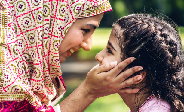 Portrait of happy lovely family arabic muslim mother and little muslim girls child with hijab dress smiling and having fun kissing together in summer park stock photo