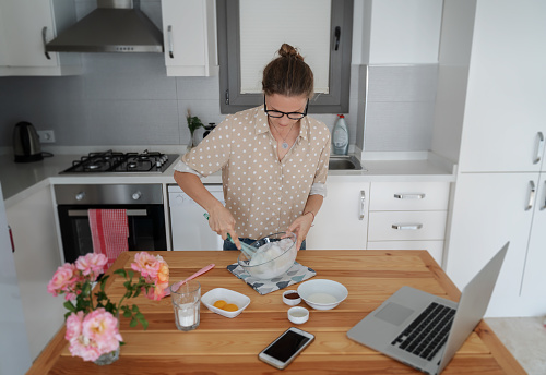 Young woman in the kitchen preparing dessert and watching the recipe on the laptop on the Internet, cooking at home, hobbies and entertainment, top view, stirring the dough with a culinary spatula