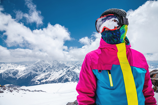 Portrait of a girl skier in a colorful bright jacket in a ski mask with a covered face on a sunny day. against the background of snow-capped mountains and clouds.