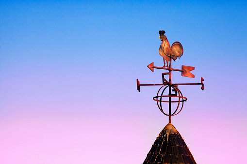 Close up of weathervane in rooster shape silhouetted on a rooftop, sunset sky in the background with copy space on the left. A Coruna province , Galicia, Spain.