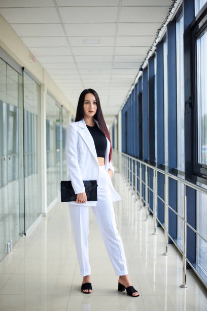 Full length portrait of content young pretty Indian business woman looking at camera, standing on one leg and leaning against wall with arms crossed. Isolated side view on white background.
