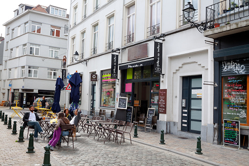 Some people are sitting outside of cafe and bar in Brussels city in street in district Ixelles near Matonge and square Place Boniface. A child is leaning at woman. A woman is standing on balcony in background. In street are several cafes and bars and restaurants