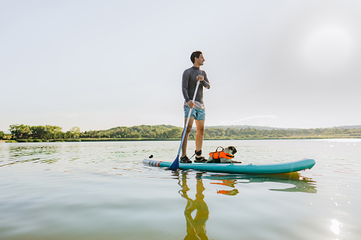Photo of a young man and his dog stand-up paddleboarding on the lake; enjoying the beautiful, warm summer afternoon, far from the hustle of the city.