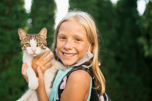 Portrait of a young little Caucasian girl with a protective face mask, holding a domestic cat in her arms and is going to school on a sunny day, smiling