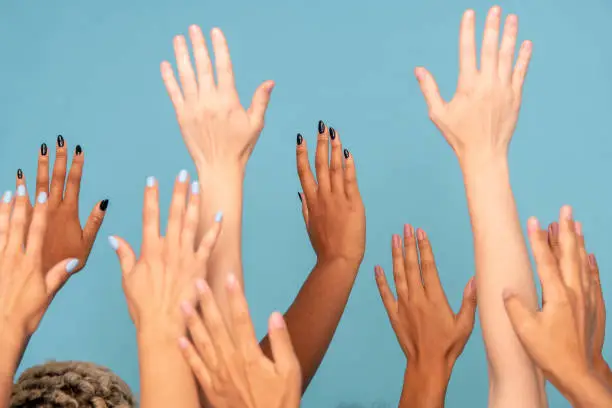 Hands of large group of young women of various ethnicities with white and dark skin raising arms in front of camera against blue background