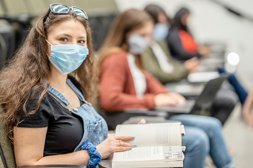 Multi-ethnic group of students wearing protective face masks while sitting in a lecture hall sitting 2 meters apart.