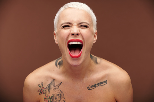 Young masculine female with short blond hair, red lipstick on lips and tattoos on chest and neck shouting and expressing aggression