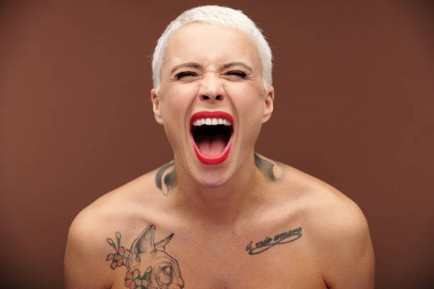 Female with short blond hair, red lipstick on lips and tattoos on chest and neck Young masculine female with short blond hair, red lipstick on lips and tattoos on chest and neck shouting and expressing aggression mouth open stock pictures, royalty-free photos & images