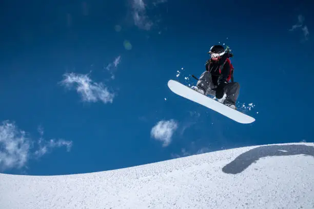 Photo of A professional snowboarder girl in flight after jumping from a snow eaves makes a rake against blue sky in sunny day. The concept of winter sports freeride and freestyle