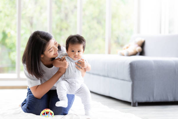 szczęśliwe małe dziecko uczy się chodzić z pomocą matki w salonie. dziecko stawia pierwsze kroki z pomocą matki i wsparcie z miłością. - family walking child toddler zdjęcia i obrazy z banku zdjęć