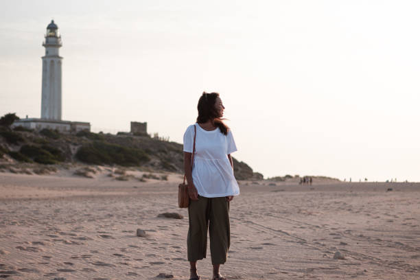 istock Young brown woman watching a sunset on a beach with a lighthouse behind her. 1267705014
