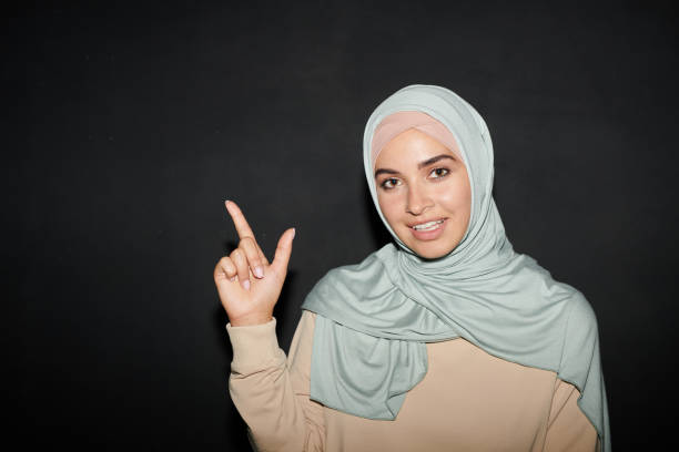 красивая молодая женщина ближнего востока носить хиджаб указывая пальцем на что-то глядя на камеру улыбается, черный фон - women middle eastern ethnicity islam hijab стоковые фото и изображения