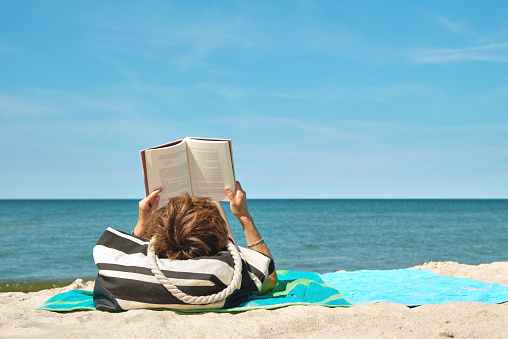 Caucasian woman lying on the beach reading a book on bright summer day