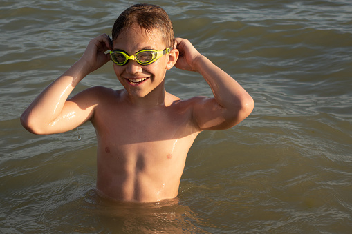 Boy 10 years old in green swimming goggles bathes and smiles in the open sea