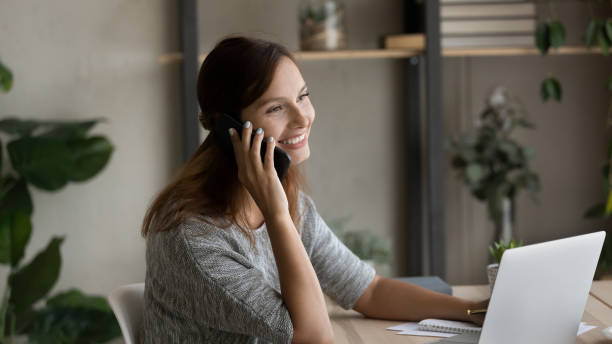 istock Smiling woman talk on smartphone working on laptop 1267719515