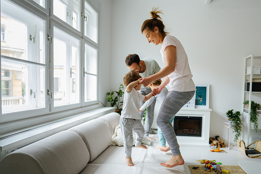 Photo of a young family jumping on the bed in the living room of their apartment; being cheerful and playful and enjoying their morning.