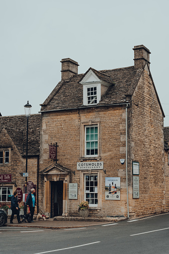 Bourton-on-the-Water, UK - July 10, 2020: Facade of Cotswolds Distillery in Bourton-on-the-Water, a famous village in rural Cotswolds area of England, couple with a dog walking past.