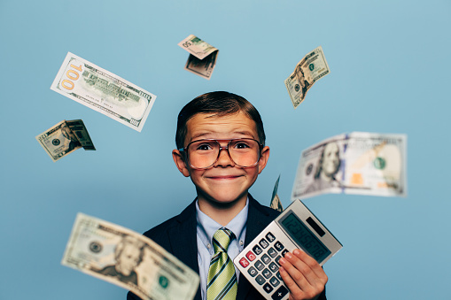 A young boy businessman wearing glasses and suit watches U.S. currency while more falls from above. He is smiling and ready to make more money as a recruit for your business.