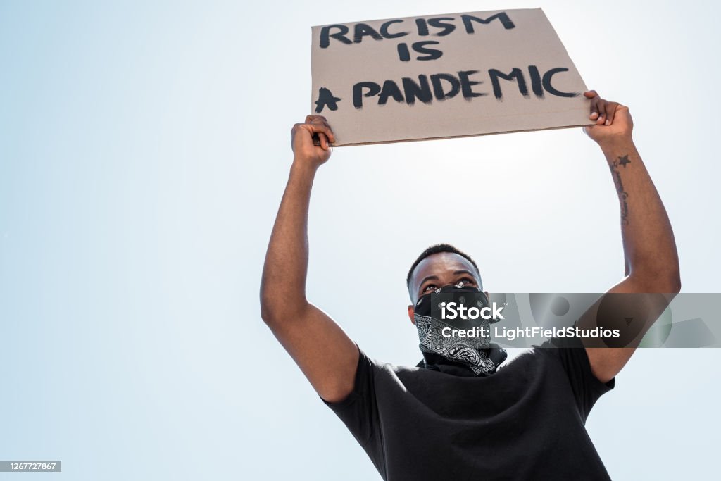 low angle view of african american man with scarf on face holding placard with racism is a pandemic lettering against blue sky Anti-racism Stock Photo